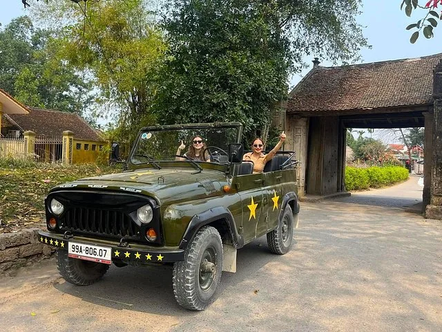 Dos mujeres viajan en un jeep militar antiguo frente a la puerta de un pueblo.